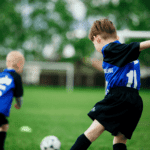 Photo of neighbourhood soccer in action. Two Highlands Rockets team members in blue and black soccer uniforms running in a green soccer field. Both kids are facing a net in the background, not the camera.