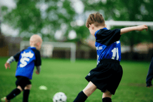 Photo of neighbourhood soccer in action. Two Highlands Rockets team members in blue and black soccer uniforms running in a green soccer field. Both kids are facing a net in the background, not the camera.